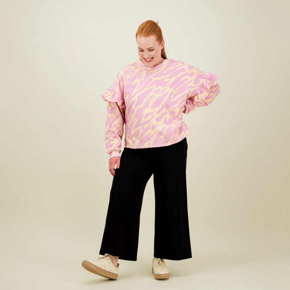 Naisten oversize-mallinen purple-sävyinen collegepaita frillahihoilla ja graafisella kuosilla, paita yhdistetty musta culottes housuihin. 